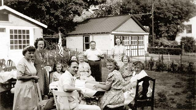 11 people ages 10-70 sit around a table in a back yard; a woman unrolls a large document on the table; 2 garages are visible behind them;