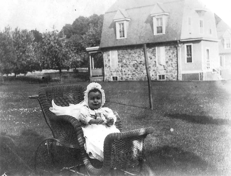 young baby dressed in a white gown and cap sits in an old fashioned pram