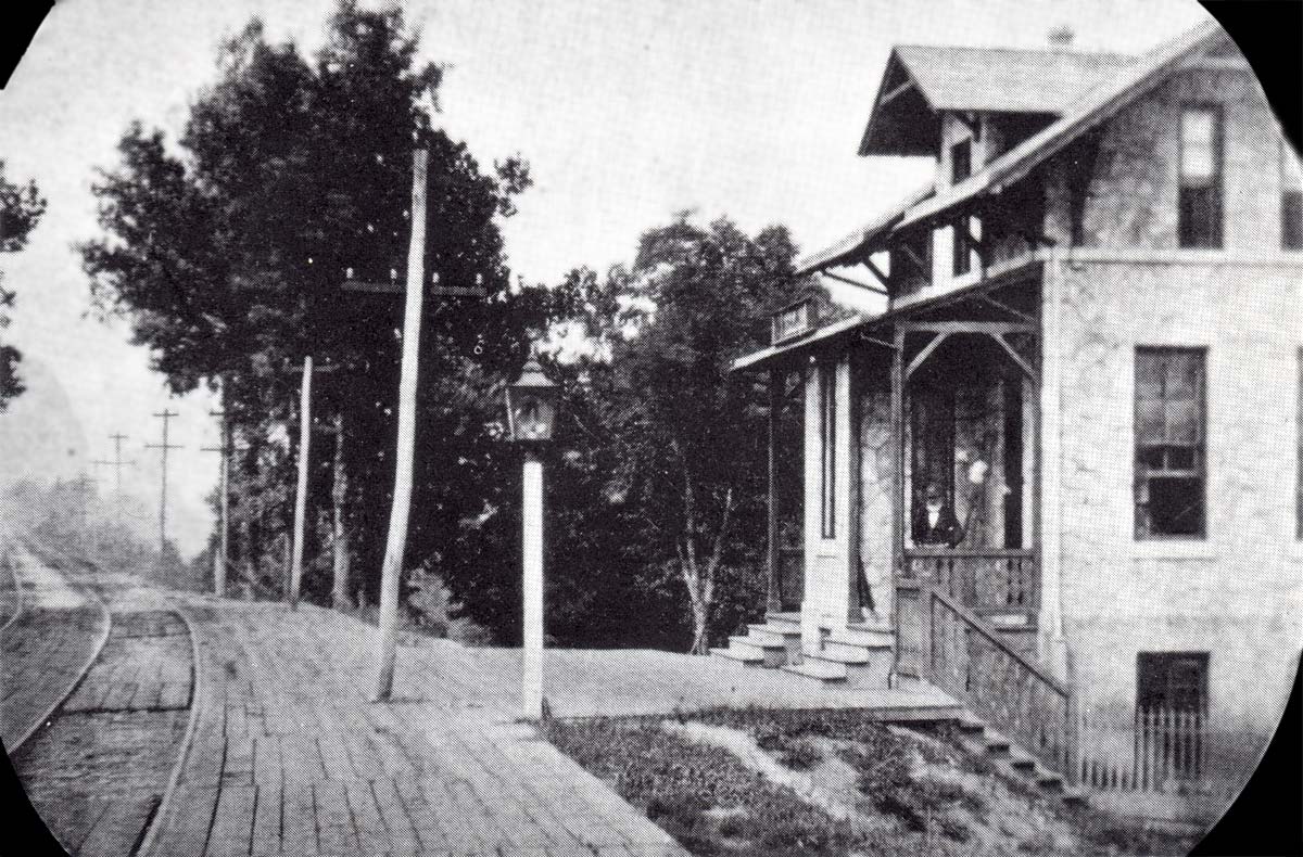 looking down a two-track rail line; beside a platform of wooden planks with a lampost and a three-story stone station house