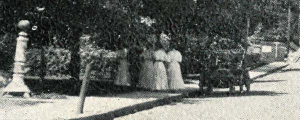 4 women in identical-looking long white dresses standing on the sidewalk under the trees; an early automobile is parked next to them.