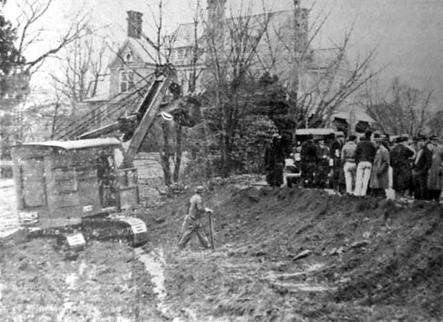 A steam shovel has gouged the earth in front of a large house; a crowd looks on.