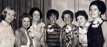seven smiling women with sculpted hairdos and flower pins