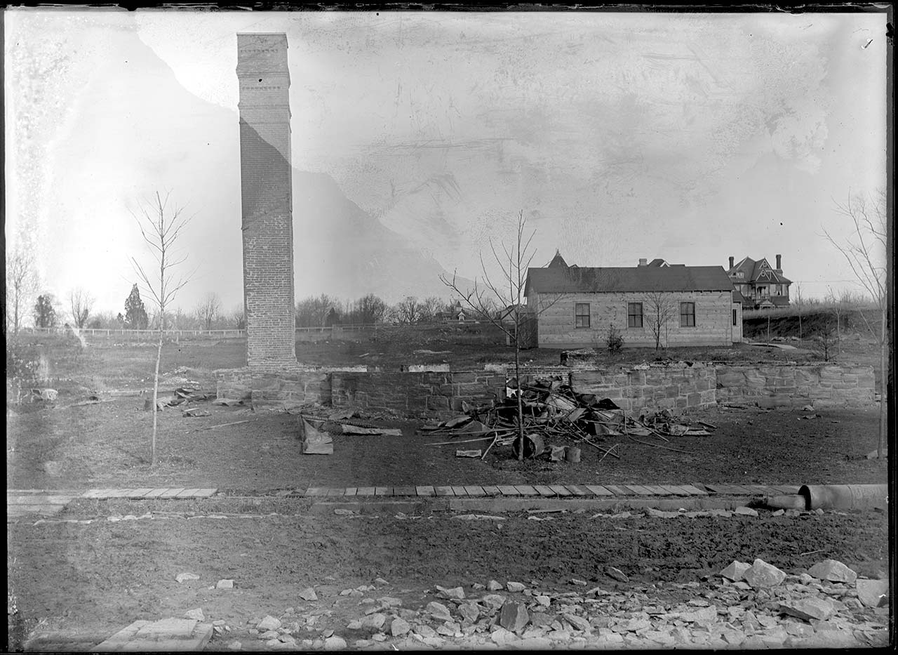 A desolate open lot with a blackened stone foundation, bare brick chimney, temporary wooden chapel behind
