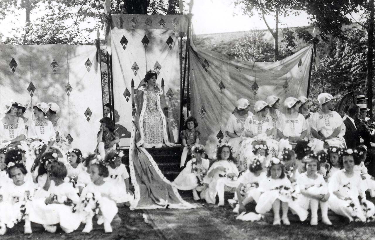 Lady Narberth in crown and long fur cape sits on a throne. Her ladies and girls in waiting sit below and around her