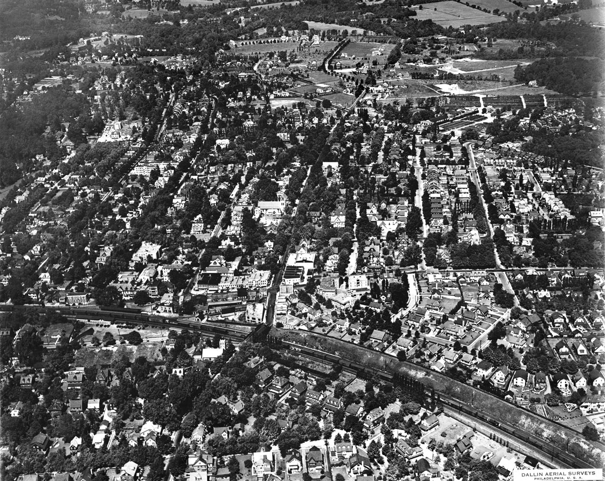An oblique low altitude aerial view of Narberth with tracks and station at bottom, receding beyond Montgomery avenue to a less developed Penn Valley.