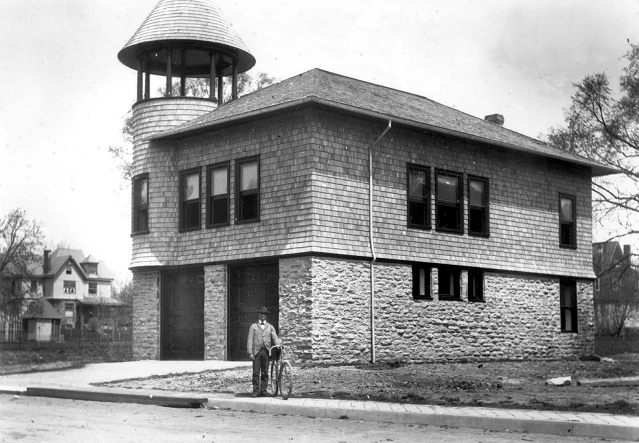 A Black man holding a bicycle stands on a paved sidewalk by a dirt street in front of a building with a stone first story and two full-height doors. A circular open watchtower capped by a pointed turret rises above the corner of its second story.