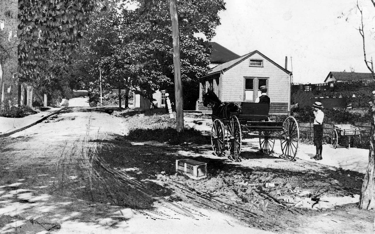 boy in knickers next to a horsecart with a seated driver, standing on a sidewalk beside a muddy lane overshaded by large leafy trees
