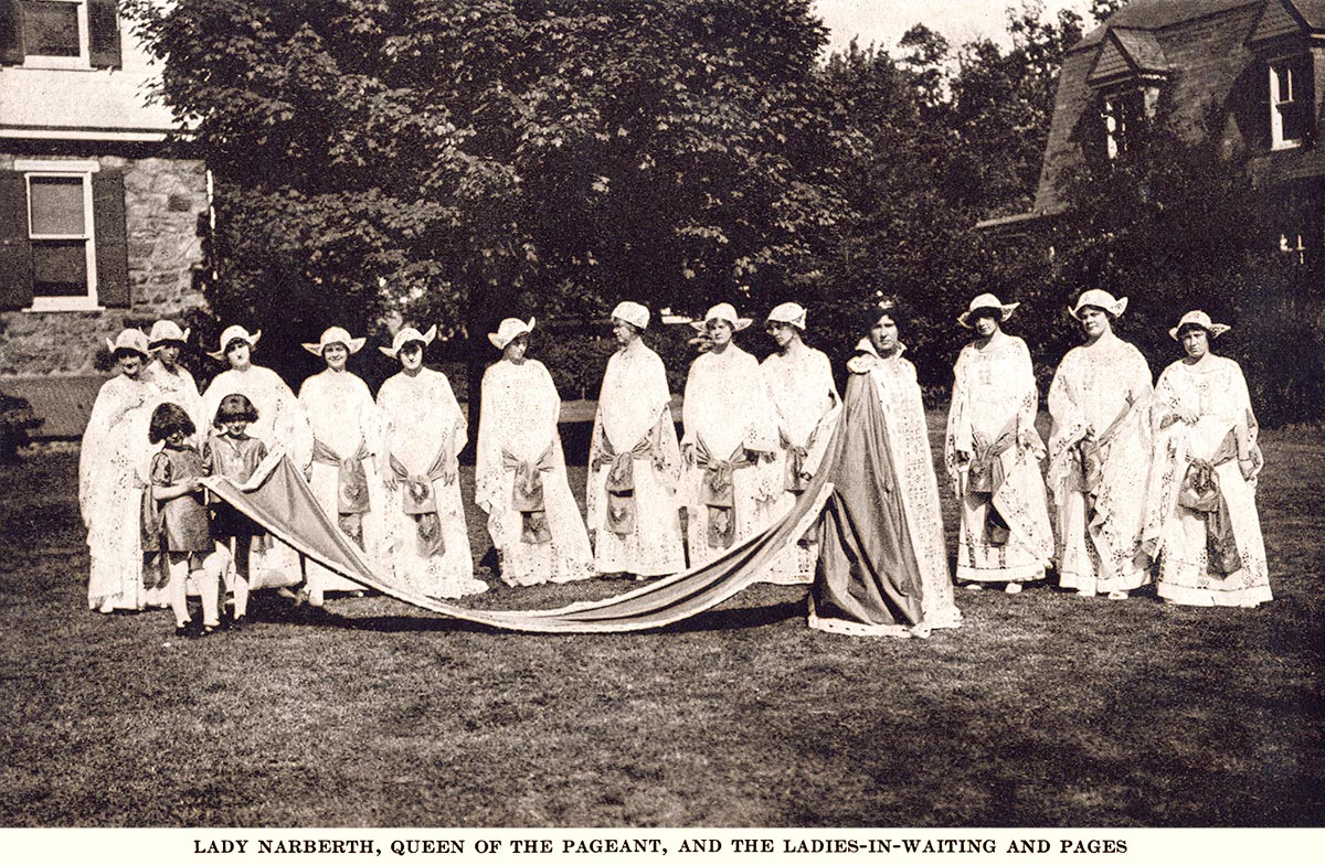 Woman wearing a crown and a long robe; two young boys in pageboy wigs hold her 10-foot cape. She is surrounded by 12 young women in elaborate matching white costumes and headdresses. They are posing in a back yard.