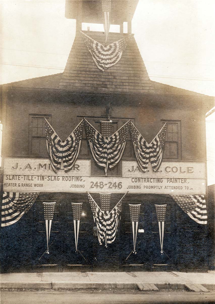 A two-story building with a roofed watchtower is festooned with stars and stripes banners on all levels. Signs painted on brick front indicate a Roofer and Painter work here.