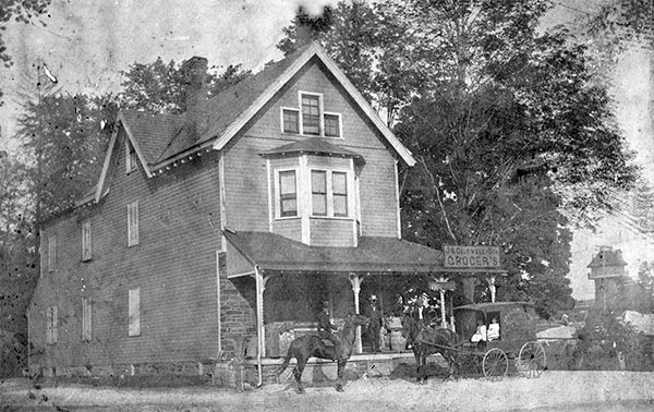A large wood frame house, 2 men stand on covered porch, a man on horseback and two children in a horse-drawn enclosed wagon in front. Signs on porch roof and wagon side read 